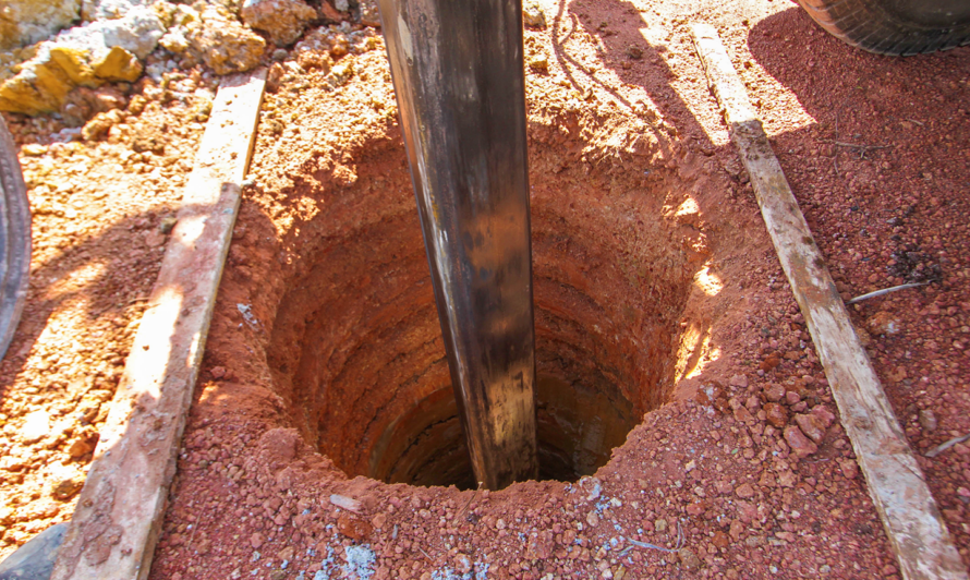 A borehole being drilled into red soil, with the shaft of a drill protruding from the hole