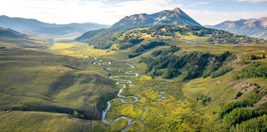 A long, windy portion of a river flowing through a blooming green mountainous landscape with clear skies