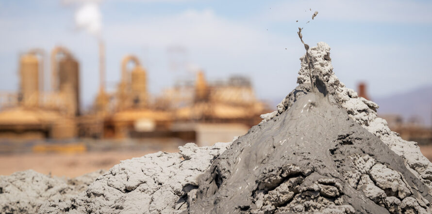 A sputtering pile of mud in the foreground spurts gray gloop into the air, while an industrial-looking power plant in the background releases steam
