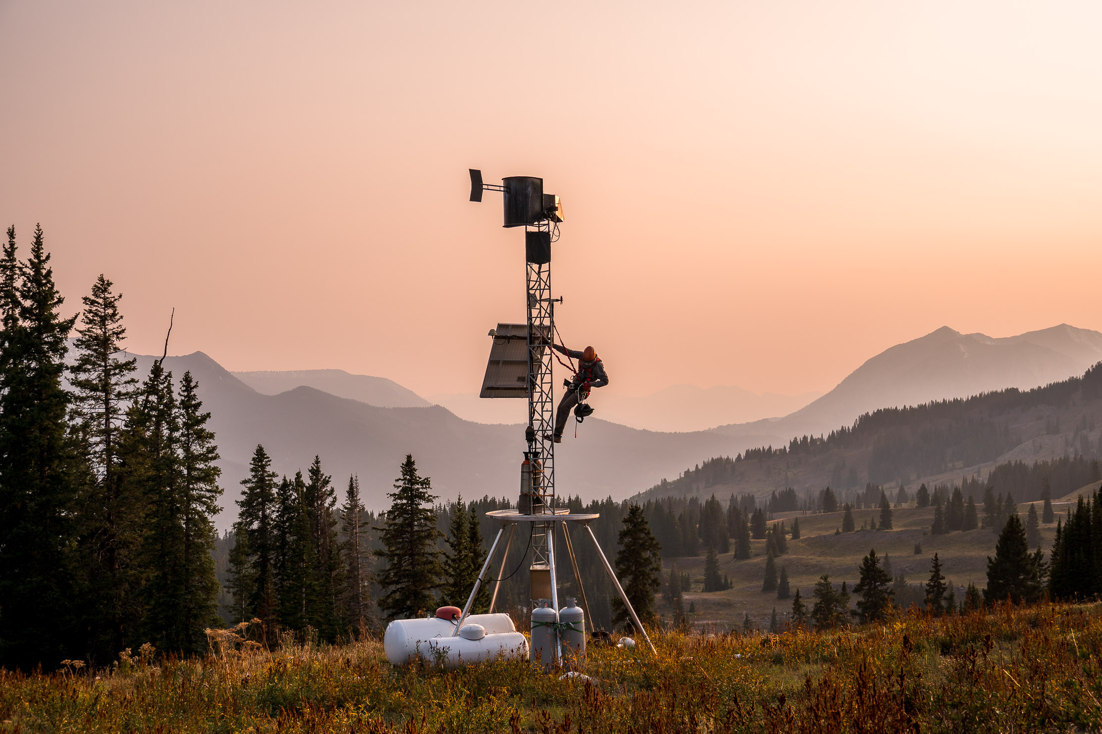 The silhouette of a lone scientist harnessed to a tall metal tower, with mountains and pine trees in the distance. The tower has instruments and devices including a solar panel and a propane flume.