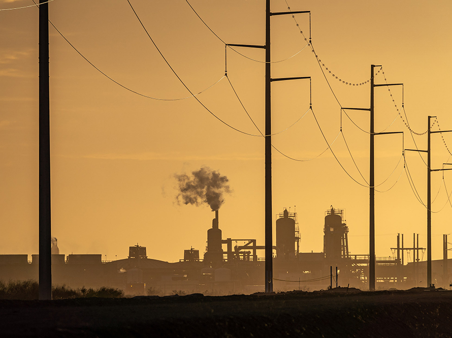 Power lines running through the foreground, while in the background a power plant releases steam on the horizon, surrounded by a light orange hazy sky