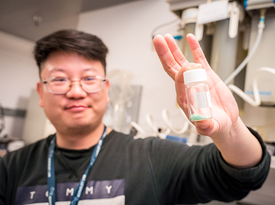 A man in glasses proudly holds up a glass vial of tiny teal beads in a scientific lab