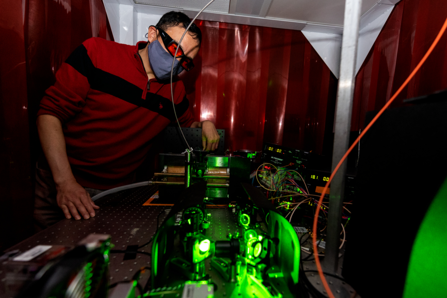 A man in a red goggles working on a complex instrument illuminated by a green laser in a dark room