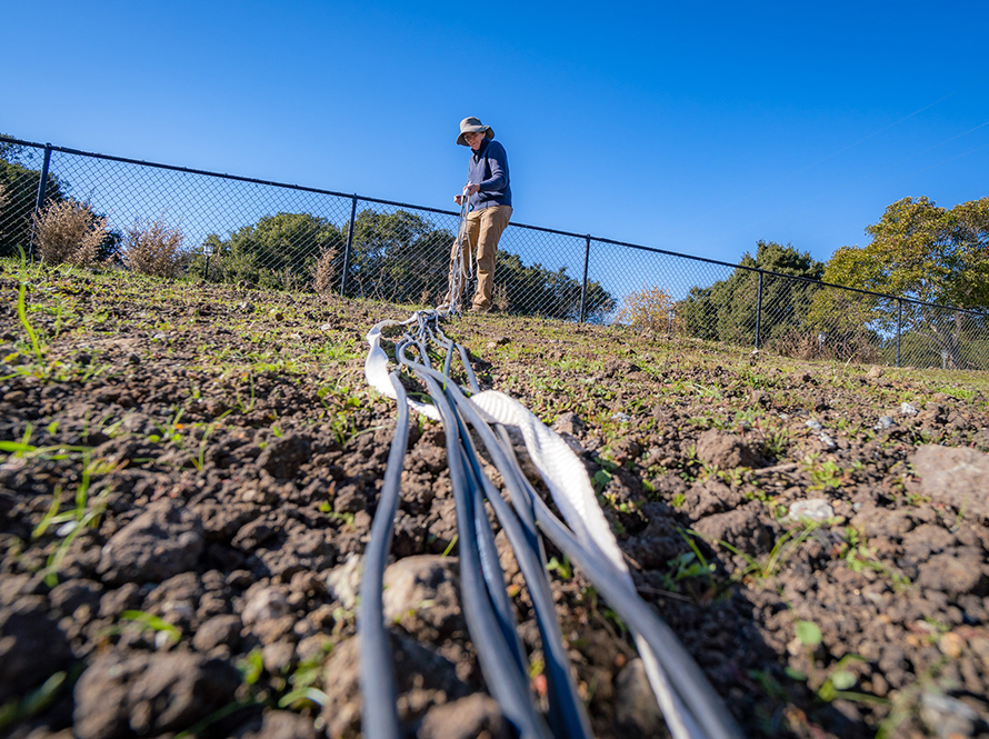 A woman stands at top of a slope in the distance with a bundle of fiber optic cables running from her hand to the foreground right in front of the camera