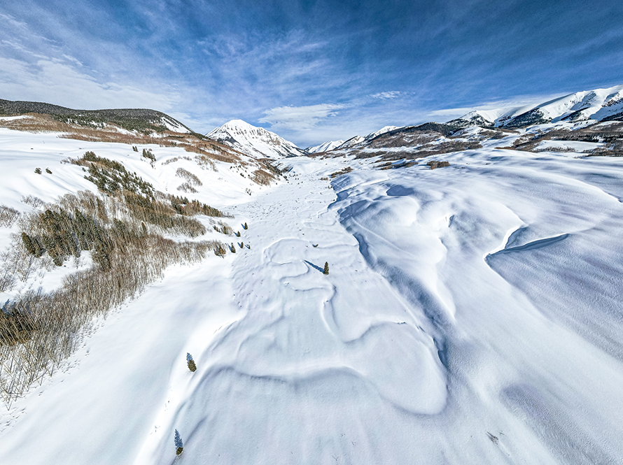 An expansive mountain valley blanketed in snow, with the trace of a river winding down the middle under the drifts