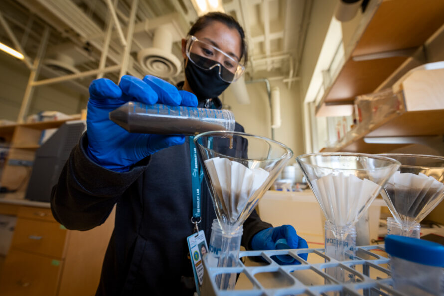 A woman in PPE pours brown liquid from a test tube into a filter