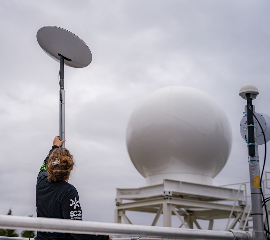 A woman on a platform holds her arm up to satellite dish equipment