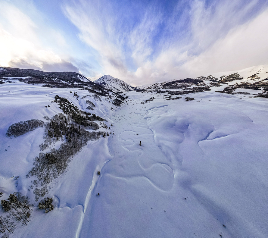 An aerial view of a snowy mountain river valley, with a dramatic peak in the distance and the winding shape of a river underneath the snow
